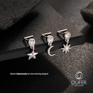 May include: Three silver nose studs with celestial charms: a star, a crescent moon, and a sun. Each stud features a clear, round gem and is displayed on a dark surface. The text "Search OuferJewelry for more stunning designs!" is visible.