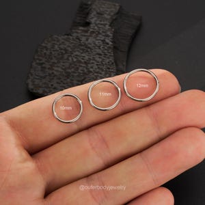 Puede incluir: Tres anillos de acero quir&uacute;rgico sin costuras de diferentes tama&ntilde;os, 10 mm, 11 mm y 12 mm, se muestran en la mano de una persona. Los anillos son ideales para piercings corporales.