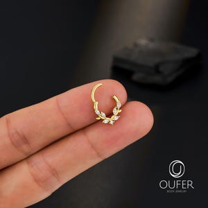 Op de afbeelding: Een goudkleurige klapcreool-oorbel met een bladvorm, versierd met heldere, ovaalvormige edelstenen. De oorbel wordt tussen vingers tegen een donkere achtergrond gehouden. Het merklogo "OUFER BODY JEWELRY" is zichtbaar.