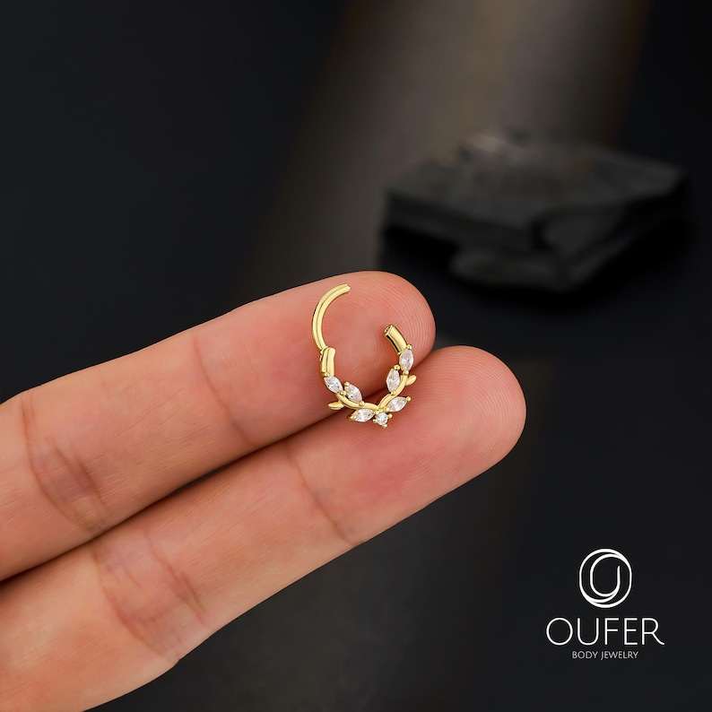 Op de afbeelding: Een goudkleurige klapcreool-oorbel met een bladvorm, versierd met heldere, ovaalvormige edelstenen. De oorbel wordt tussen vingers tegen een donkere achtergrond gehouden. Het merklogo "OUFER BODY JEWELRY" is zichtbaar.