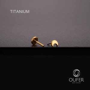 May include: Gold-coloured titanium body jewellery. One piece is a flat-backed stud, the other is a crescent moon shape. The word "TITANIUM" is printed in white at the top of the image. The jewellery is on a dark surface.