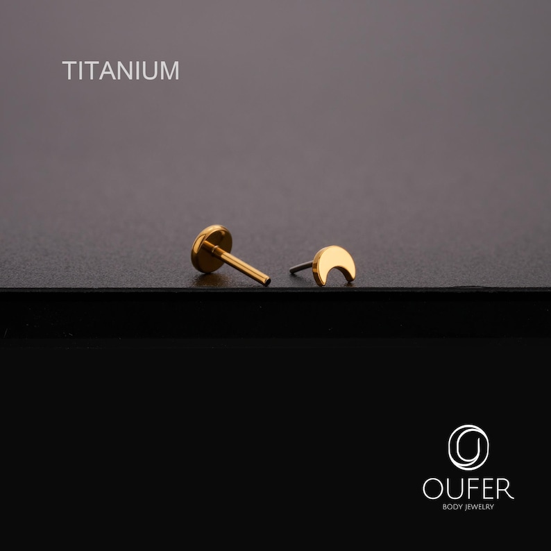 May include: Gold-coloured titanium body jewellery. One piece is a flat-backed stud, the other is a crescent moon shape. The word "TITANIUM" is printed in white at the top of the image. The jewellery is on a dark surface.