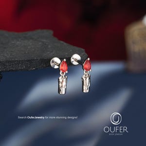 May include: A pair of silver-toned stud earrings featuring a dangling charm shaped like a melting candle with a red teardrop-shaped gemstone at the top. The text "Search OuferJewelry for more stunning designs!" and the "OUFER BODY JEWELRY" logo are visible.