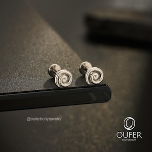 May include: A pair of silver-toned metal stud earrings featuring a spiral swirl design. Each earring has a flat circular back post and a front face shaped like a coiled spiral. The text @ouferbodyjewelry and the OUFER BODY JEWELRY logo are visible.