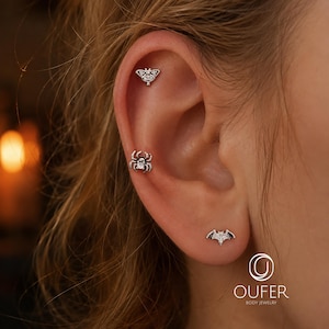 May include: Three silver-toned metal stud earrings shaped like a moth, a spider, and a bat. The moth is positioned at the top of the ear cartilage, the spider is in the middle, and the bat is on the earlobe. Each piece features small clear stones.