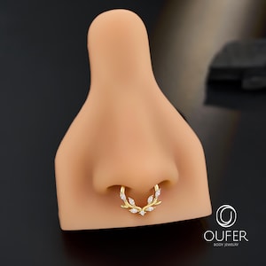 Op de afbeelding: Een gouden septumring met een bladvorm, versierd met kleine, heldere edelstenen. De sieraden worden getoond op een neusmodel, waarbij de vorm en stijl worden benadrukt. De ring heeft een delicate, elegante uitstraling.