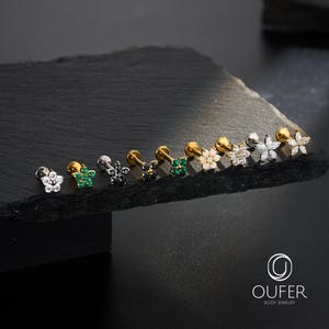 Puede incluir: Una variedad de pendientes de joyer&iacute;a corporal con forma de flor en varios colores y dise&ntilde;os. Los pendientes se exhiben sobre una superficie oscura y texturizada. La joyer&iacute;a presenta metal dorado, plateado y negro con piedras preciosas transparentes, verdes y blancas. El logotipo de la marca "OUFER BODY JEWELRY" es visible.