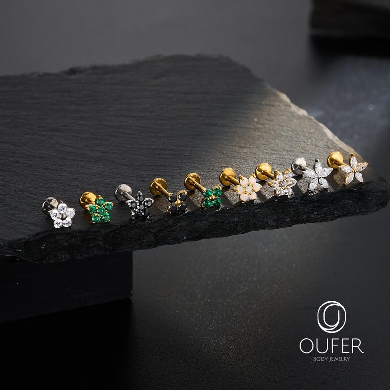 Puede incluir: Una variedad de pendientes de joyer&iacute;a corporal con forma de flor en varios colores y dise&ntilde;os. Los pendientes se exhiben sobre una superficie oscura y texturizada. La joyer&iacute;a presenta metal dorado, plateado y negro con piedras preciosas transparentes, verdes y blancas. El logotipo de la marca "OUFER BODY JEWELRY" es visible.