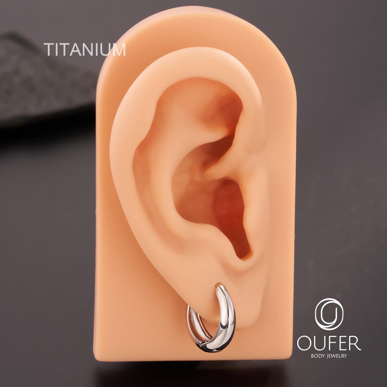 Puede incluir: Un pendiente de aro plateado se muestra en un modelo de oreja color carne. La palabra "TITANIUM" est&aacute; impresa en blanco en la parte superior izquierda. El pendiente tiene un dise&ntilde;o simple y curvado, que refleja la luz. El logotipo de OUFER Body Jewelry es visible.