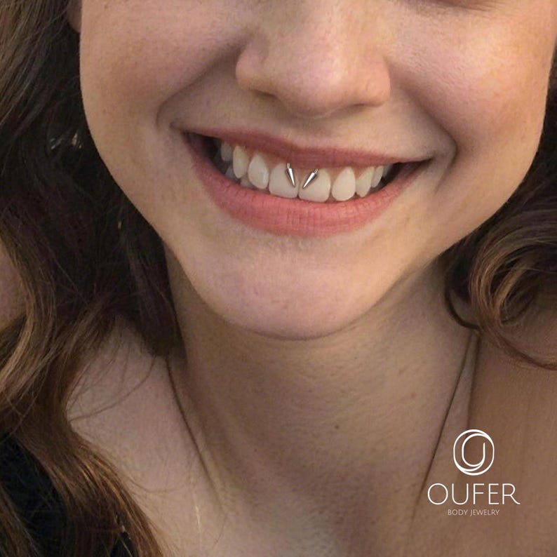May include: A silver-toned metal smiley piercing jewelry piece featuring two small, pointed spikes positioned between the upper teeth. The jewelry is displayed in a mouth with the OUFER BODY JEWELRY logo visible in the bottom right corner.
