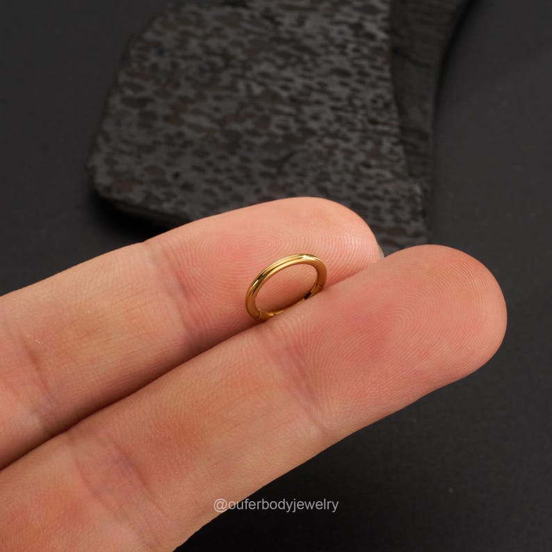 May include: A gold-colored seamless ring, also known as a captive bead ring, is shown on a finger. The ring is a popular choice for body piercings.