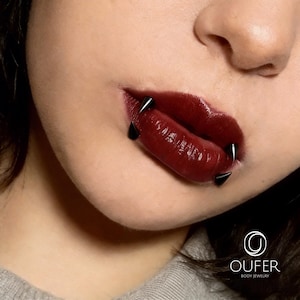 May include: Four black spike-shaped lip piercings are positioned around the mouth. The jewelry features sharp, conical ends that extend outward from the lips. The lips are coated in a glossy, dark burgundy lipstick, contrasting with the black metal.
