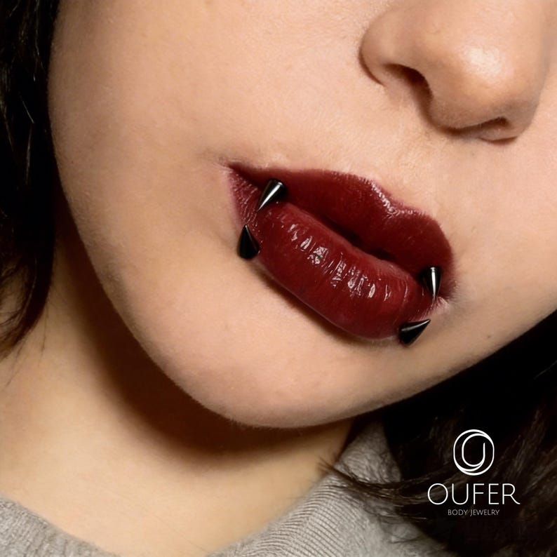 May include: Four black spike-shaped lip piercings are positioned around the mouth. The jewelry features sharp, conical ends that extend outward from the lips. The lips are coated in a glossy, dark burgundy lipstick, contrasting with the black metal.
