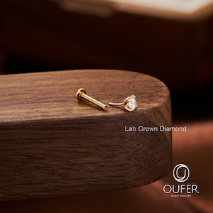 Puede incluir: Un pendiente labret dorado con un diamante transparente de talla redonda cultivado en laboratorio. El pendiente est&aacute; sobre una superficie de madera oscura. El texto "Lab Grown Diamond" es visible. El logotipo de Oufer Body Jewelry est&aacute; en la esquina inferior derecha.