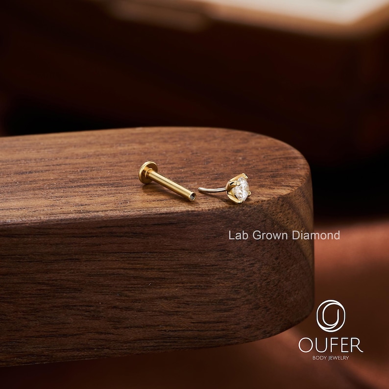 Puede incluir: Un pendiente labret dorado con un diamante transparente de talla redonda cultivado en laboratorio. El pendiente est&aacute; sobre una superficie de madera oscura. El texto "Lab Grown Diamond" es visible. El logotipo de Oufer Body Jewelry est&aacute; en la esquina inferior derecha.