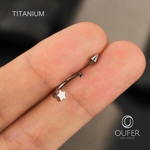 May include: A silver-colored titanium curved barbell body jewelry. The jewelry features a star-shaped end and a pointed cone-shaped end. The word "TITANIUM" is printed in white on the top left of the image.