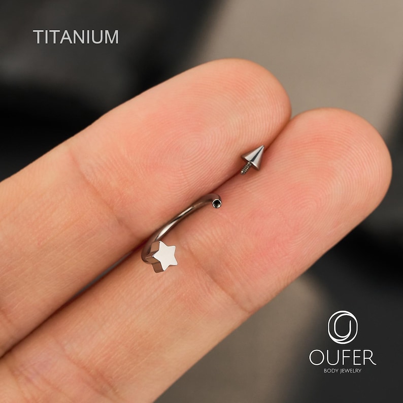 May include: A silver-colored titanium curved barbell body jewelry. The jewelry features a star-shaped end and a pointed cone-shaped end. The word "TITANIUM" is printed in white on the top left of the image.