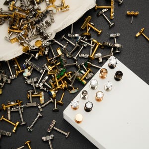 Puede incluir: Un surtido de joyas para el cuerpo, que incluye barras y expansores dorados y plateados. Algunos pendientes presentan piedras preciosas transparentes, mientras que otros tienen acentos de color. Las joyas est&aacute;n esparcidas sobre una superficie blanca sobre un fondo oscuro.