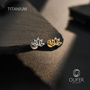 Op de afbeelding: Twee titanium oorknopjes met platte achterkant in de vorm van lotusbloemen. De ene knop is zilverkleurig met heldere steentjes, de andere is goudkleurig met heldere steentjes. De tekst TITANIUM en OUFER BODY JEWELRY is zichtbaar.