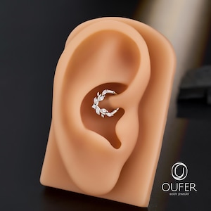 Op de afbeelding: Een zilverkleurige, bladvormige oorbel met heldere edelstenen wordt getoond in een tan oormodel. De oorbel is een oorring, met de edelstenen langs de buitenste curve gerangschikt. De merknaam "OUFER BODY JEWELRY" is zichtbaar in de rechterbenedenhoek.