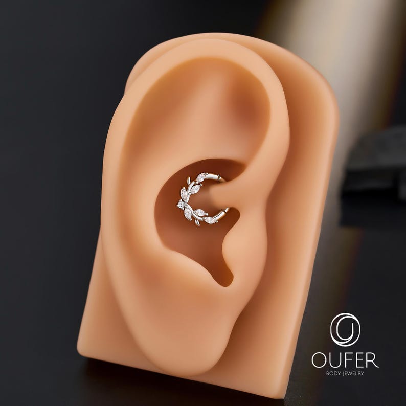 Op de afbeelding: Een zilverkleurige, bladvormige oorbel met heldere edelstenen wordt getoond in een tan oormodel. De oorbel is een oorring, met de edelstenen langs de buitenste curve gerangschikt. De merknaam "OUFER BODY JEWELRY" is zichtbaar in de rechterbenedenhoek.