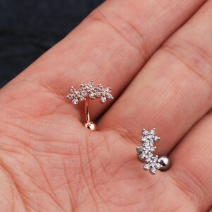 May include: Two belly button rings with a flower design. The rings are made of metal and have clear stones. One ring is rose gold and the other is silver.