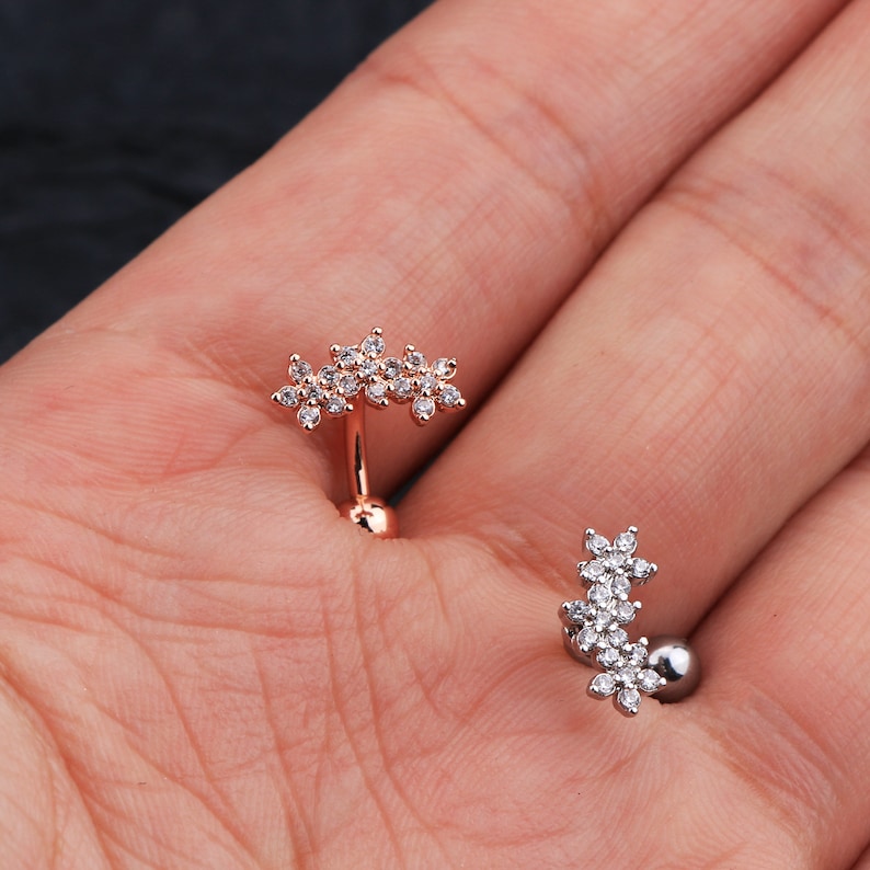 May include: Two belly button rings with a flower design. The rings are made of metal and have clear stones. One ring is rose gold and the other is silver.