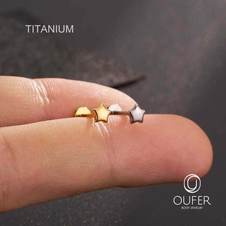 K&ouml;nnte beinhalten: Nahaufnahme eines K&ouml;rperschmuckst&uuml;cks aus Titan mit drei sternf&ouml;rmigen Nieten in Gold, Silber und Gold. Das Wort "TITANIUM" ist oben in Wei&szlig; aufgedruckt. Der Schmuck wird zwischen zwei Fingern gehalten.