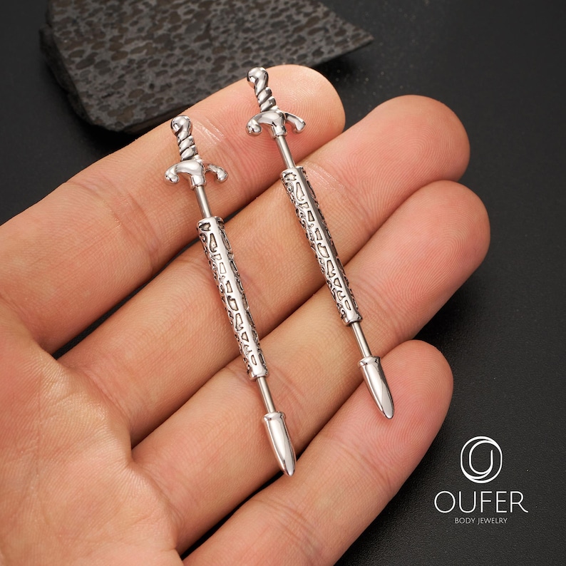 K&ouml;nnte beinhalten: Zwei silberfarbene, schwertf&ouml;rmige K&ouml;rperschmuckst&uuml;cke. Jedes St&uuml;ck hat einen detaillierten Knauf, eine strukturierte Klinge und eine spitze Spitze. Der Schmuck wird in einer Hand vor dunklem Hintergrund gehalten. Das Markenlogo "OUFER BODY JEWELRY" ist sichtbar.