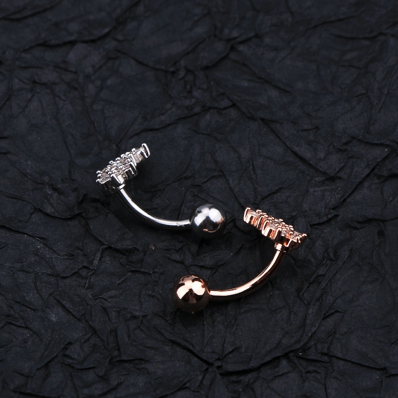 May include: Two belly button rings with a jeweled design. One ring has a silver ball and the other has a rose gold ball.
