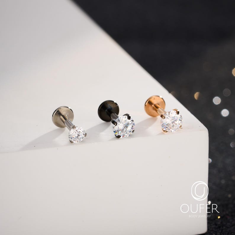 Puede incluir: Tres pendientes de bot&oacute;n con piedras de circonita c&uacute;bica redondas y transparentes. Los pendientes tienen acabados plateados, negros y oro rosa. Los pendientes se exhiben sobre una superficie blanca con un fondo borroso.