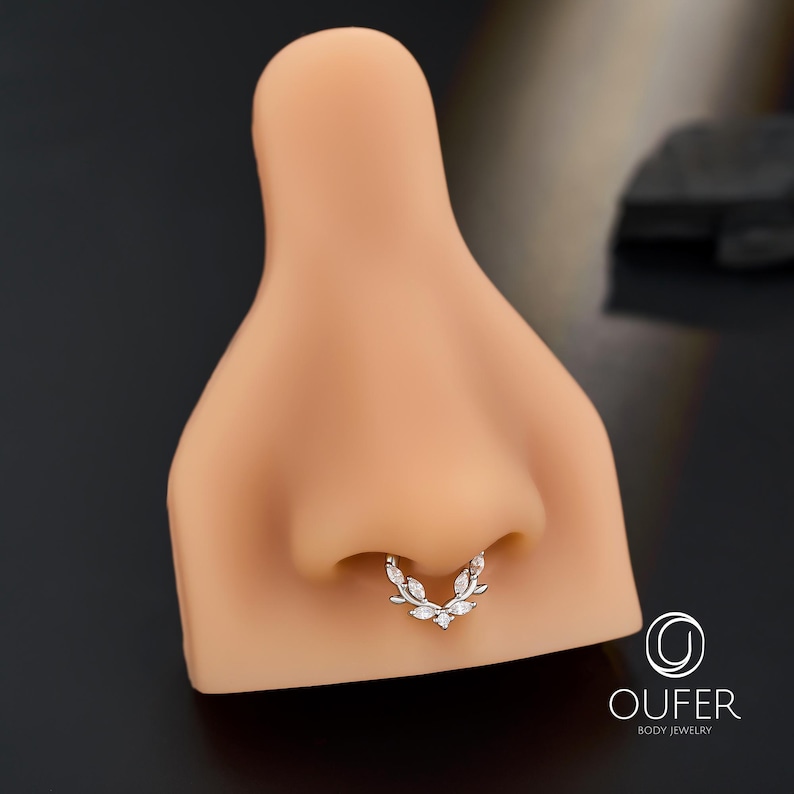 Op de afbeelding: Een zilveren septumring met een bladvorm, versierd met heldere edelstenen. De sieraden worden getoond op een neusmodel, waarbij de vorm en stijl worden benadrukt. Het merklogo "OUFER BODY JEWELRY" is zichtbaar.