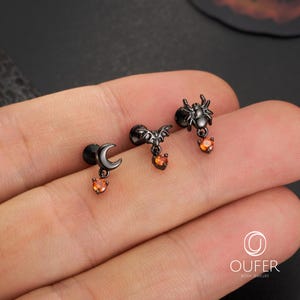 May include: Three black Halloween-themed earrings. One features a crescent moon, another a bat, and the third a spider. Each earring has a small orange heart-shaped gem. The earrings are displayed on a hand.