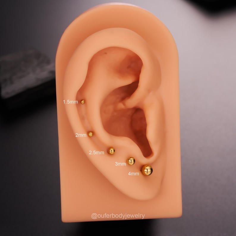 May include: Close-up of a model ear with gold ball earrings of different sizes. The earrings are labeled with their size in millimeters: 1.5mm, 2mm, 2.5mm, 3mm, and 4mm. The text "@outerbodyjewelry" is at the bottom of the image.