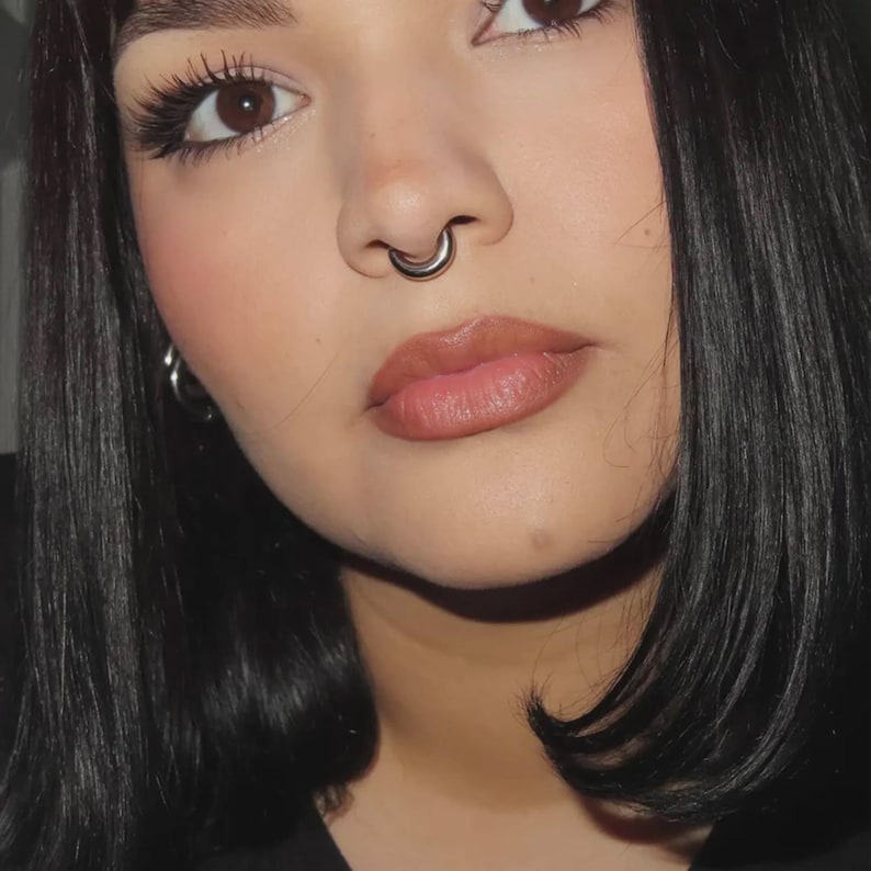 Puede incluir: Primer plano del rostro de una persona con un piercing de nariz plateado y pendientes de aro. La persona tiene cabello oscuro, ojos marrones y lleva maquillaje. El enfoque est&aacute; en los rasgos faciales y las joyas.