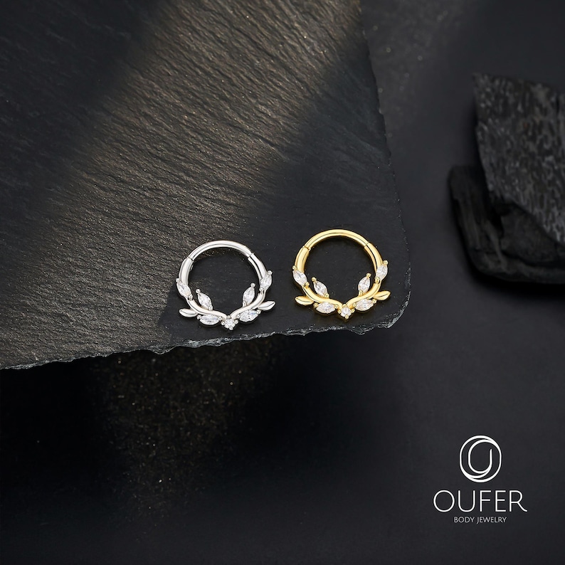 Op de afbeelding: Twee lichaamsjuwelen ringen, &eacute;&eacute;n zilver en &eacute;&eacute;n goud, elk met een bladvormgeving en heldere edelstenen. De ringen staan tegen een donkere, getextureerde achtergrond, met het merklogo rechtsonder.