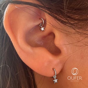 Può includere: Due orecchini a cerchio color argento con ciondoli a forma di stella, ognuno con una piccola pietra trasparente al centro. Un cerchio è posizionato nella cartilagine e l'altro nel lobo. Il testo OUFER BODY JEWELRY è visibile.