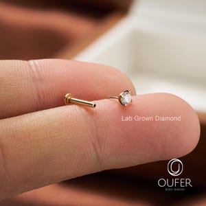 Puede incluir: Dos piezas de joyer&iacute;a corporal. Una es un labret de color dorado con un peque&ntilde;o diamante transparente. La otra es un poste de color dorado. El texto "Lab Grown Diamond" es visible. La joyer&iacute;a est&aacute; en un dedo.