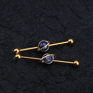 Puede incluir: Dos barras industriales doradas. Cada barra presenta un dise&ntilde;o de planeta azul con un anillo dorado y extremos de bola dorados. Los planetas tienen una apariencia moteada y estrellada. La joyer&iacute;a est&aacute; colocada sobre un fondo oscuro y texturizado.
