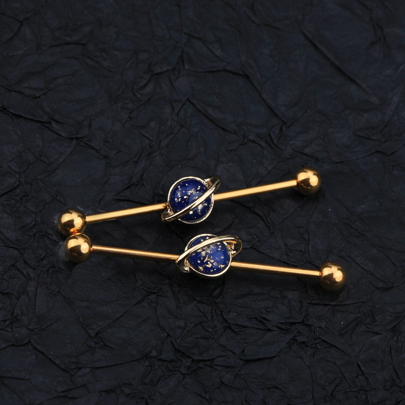 Puede incluir: Dos barras industriales doradas. Cada barra presenta un dise&ntilde;o de planeta azul con un anillo dorado y extremos de bola dorados. Los planetas tienen una apariencia moteada y estrellada. La joyer&iacute;a est&aacute; colocada sobre un fondo oscuro y texturizado.