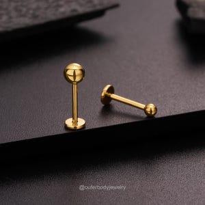 May include: Two gold-colored barbell earrings with round balls on each end. The earrings are on a black surface.