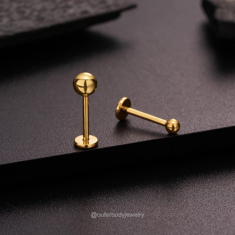 May include: Two gold-colored barbell earrings with round balls on each end. The earrings are on a black surface.
