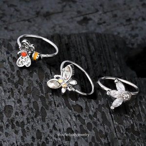 Op de afbeelding: Drie zilveren neusringen met verschillende bedels. De eerste ring heeft een bij-bedel met gele en oranje accenten. De tweede ring heeft een vlinderbedel met heldere strass steentjes. De derde ring heeft een bloembedel met heldere strass steentjes.