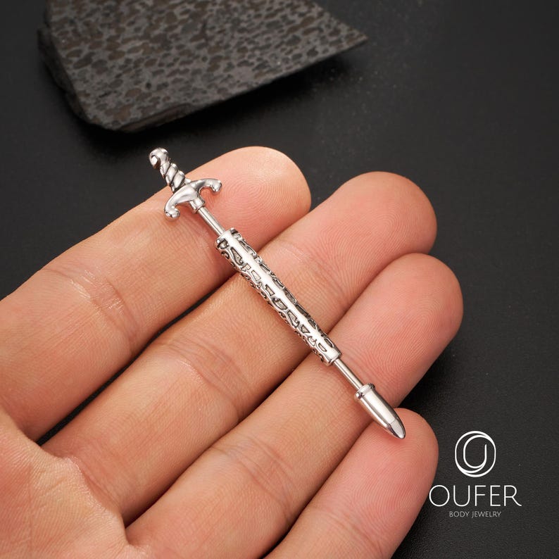 K&ouml;nnte beinhalten: Ein silberfarbenes K&ouml;rperschmuckst&uuml;ck in Form eines Schwertes. Das Schwert hat ein detailliertes Griff- und Klingendesign. Der Schmuck wird in einer Hand vor dunklem Hintergrund gehalten. Der Markenname "OUFER BODY JEWELRY" ist sichtbar.