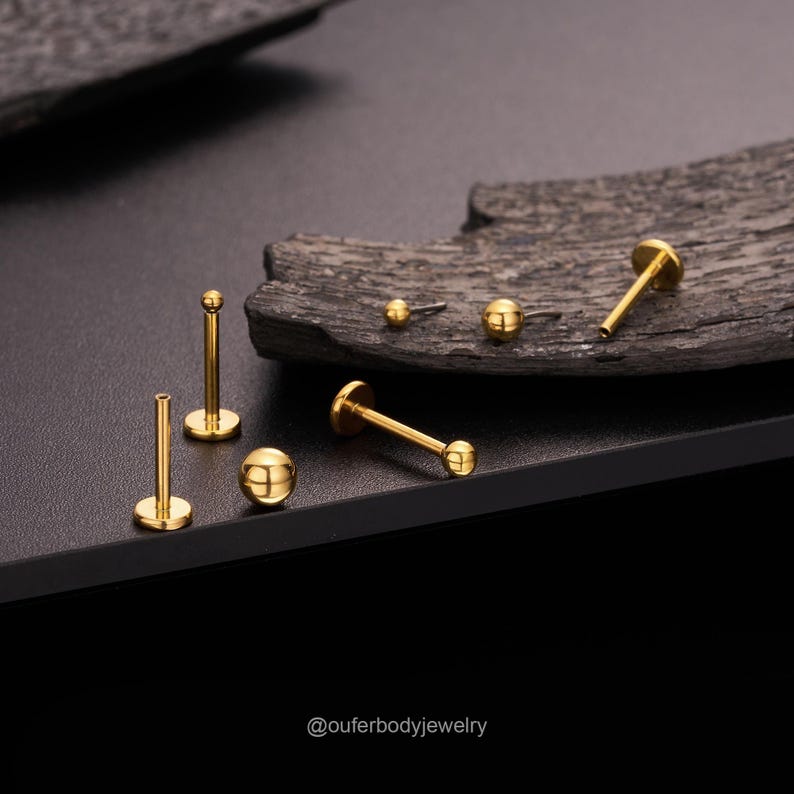 May include: A set of six gold-colored body jewelry pieces, including labret studs and a ball, on a dark wooden surface. The jewelry is arranged in a semi-circle. The text '@outerbodyjewelry' is visible in the bottom right corner of the image.