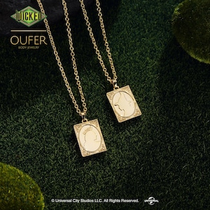 May include: Two gold-toned necklaces featuring rectangular pendants with engraved silhouettes of figures in profile. The pendants have ornate borders and are suspended from delicate gold chains. Text includes WICKED, OUFER BODY JEWELRY, and Universal City Studios LLC. All Rights Reserved.