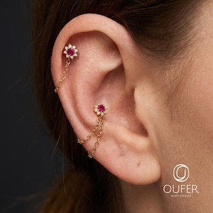 May include: Gold ear jewelry featuring two flower-shaped studs with central red stones and surrounding clear stones. One stud has a single gold chain, while the other has two gold chains connecting to the post. Text reads OUFER BODY JEWELRY.