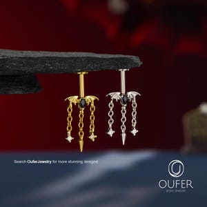 Può includere: Due piercing per l'ombelico, uno color oro e uno color argento, con un design ad ali di pipistrello, una pietra nera taglio marquise, tre catenelle pendenti e piccole pietre trasparenti. Il testo "Search OuferJewelry for more stunning designs!" è visibile.