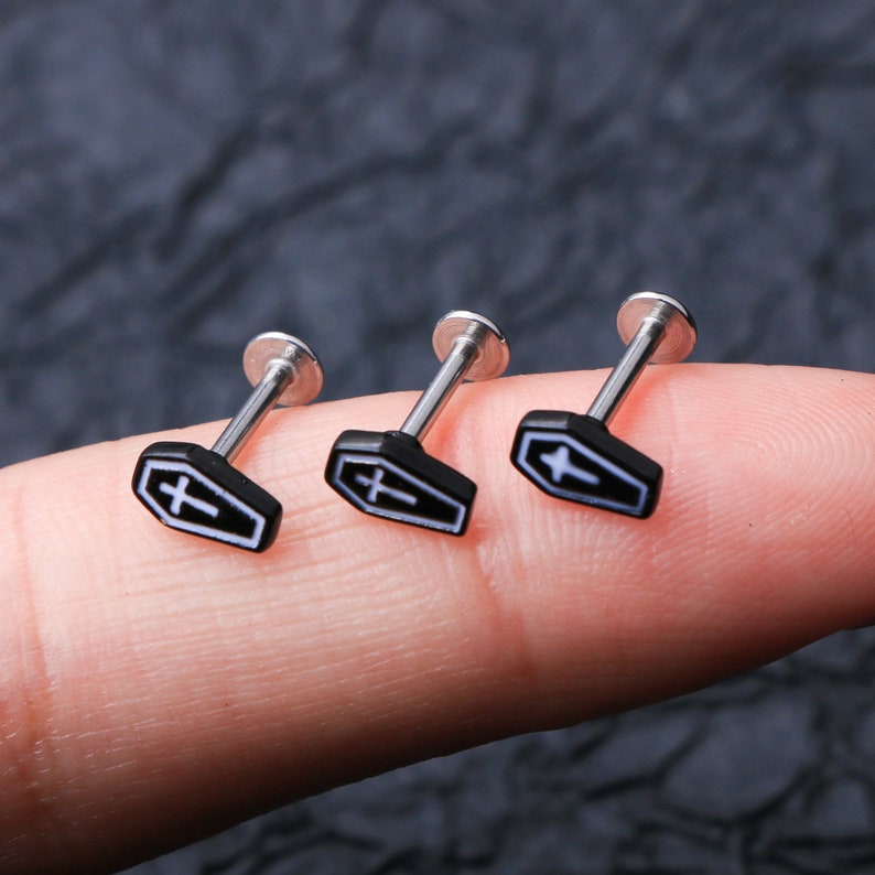 May include: Three silver body piercing studs with black coffin-shaped charms featuring a white cross.