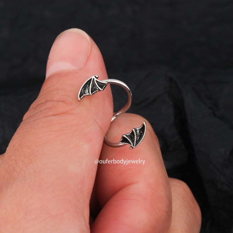Peut inclure: Bague en argent avec deux motifs d'ailes de chauve-souris noires. La bague est port&eacute;e sur un doigt.